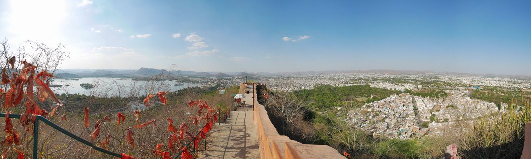 Panoramic photograph showing Udaipur city with white buildings, Lake Pichola water body, red prayer flags on bushes in foreground, stone pathway with railing, and Aravalli mountains in the distance under blue sky