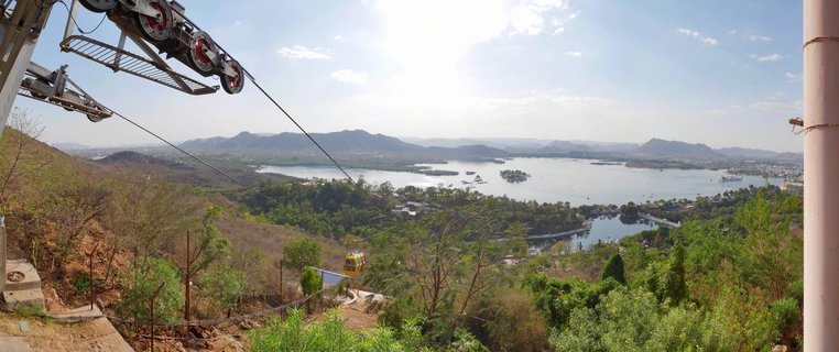 Cable car machinery and yellow gondola suspended on cables overlooking a large lake with islands, surrounded by hills and vegetation under a bright sky in Udaipur, India
