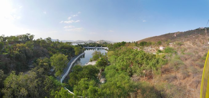 Panoramic landscape showing a lake winding through green vegetation with hills on both sides and palace buildings visible in the distance under blue sky