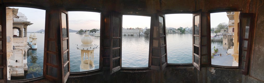 View of Lake Pichola with Taj Lake Palace visible through five open wooden windows from an interior stone room