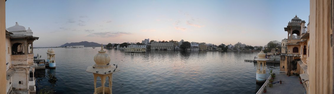 Panoramic view of Lake Palace on Lake Pichola at sunset in Udaipur, with traditional Rajasthani domed pavilions in the foreground and the Aravalli Mountains in the background