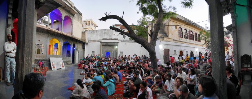 Large crowd of people sitting on red carpets in a traditional Indian courtyard surrounded by cream-colored buildings with arched doorways lit in blue, purple, and yellow colors, with a large tree in the center