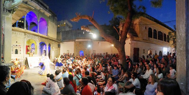 Traditional Rajasthani folk dancers in blue and white costumes perform in a historic courtyard at night while musicians play and a large audience watches