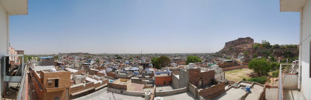Wide panoramic cityscape of Jodhpur showing densely packed buildings with flat rooftops and Mehrangarh Fort on a rocky hill in the background under clear blue sky