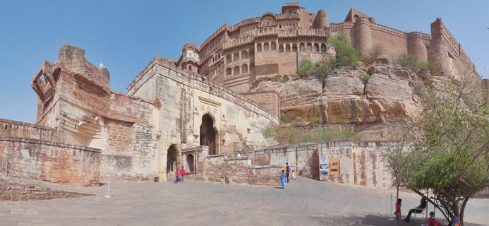 Wide view of Mehrangarh Fort in Jodhpur showing the entrance courtyard with tourists walking toward arched gateways, with the massive sandstone fortress rising on cliffs behind, featuring multiple levels of palaces and defensive walls under a clear blue sky