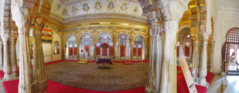 Interior view of ornate palace hall in Jodhpur with gilded decorative columns, painted ceiling with portrait medallions, arched stained-glass windows, and red carpet flooring