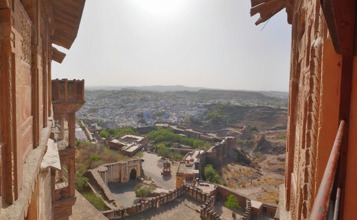 View from Mehrangarh Fort in Jodhpur, India, showing red sandstone fortress walls and windows framing the famous Blue City with its indigo-painted traditional houses stretching across the arid Rajasthan landscape under a hazy sky
