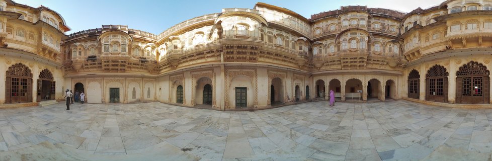 Wide courtyard of historic Indian palace with ornate sandstone balconies, carved archways, and visitors walking on marble flooring under blue sky