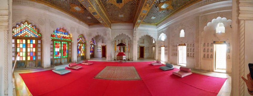 Interior of ornate Indian palace hall with white carved walls, colorful stained glass windows, red carpet, floor cushions, and decorated ceiling