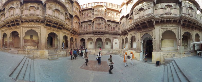 Panoramic view of an ornate sandstone courtyard at Mehrangarh Fort with carved balconies, arched doorways, and visitors walking on stone pavement