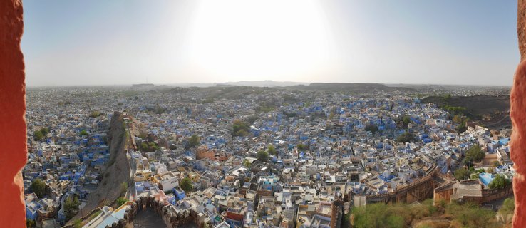 Panoramic view of densely packed blue-painted houses in Jodhpur's old city, framed by red sandstone fort walls, with arid hills visible in the distance under a bright clear sky