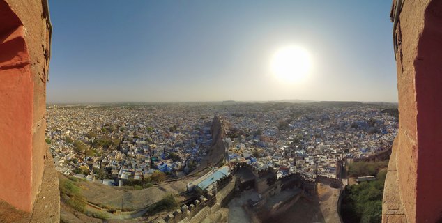 Panoramic view of Jodhpur's blue houses and city fortifications seen through red sandstone fort walls under bright sun and clear blue sky