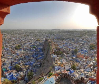 Panoramic view of densely packed blue-painted buildings in Jodhpur, India, framed by a red sandstone archway with fortification walls running along a rocky ridge through the city