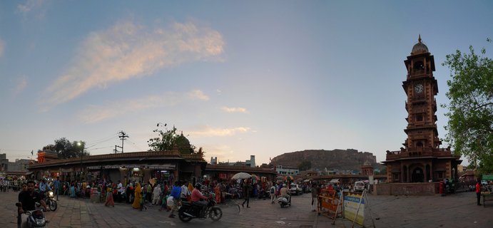 A tall red sandstone clock tower rises above a busy market square in Jodhpur, India, with people walking and riding motorcycles across stone pavement, shops lining the left side, and a fort visible on a hill in the distance under a pale blue evening sky with scattered clouds