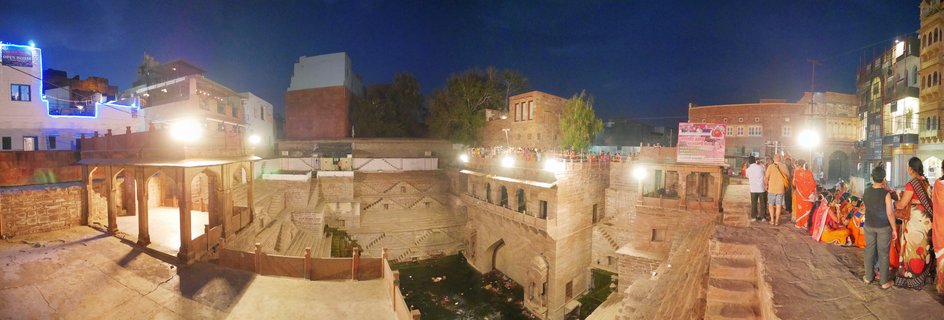 Nighttime panoramic view of a historic Indian stepwell with illuminated sandstone architecture, geometric staircases, and people in colorful traditional clothing gathered on terraces