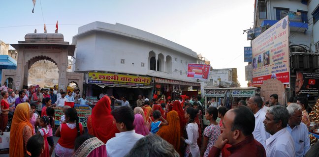 A crowded street scene in Pushkar, India showing pilgrims in colorful traditional Indian clothing walking past shops with Hindi signs near a temple entrance archway