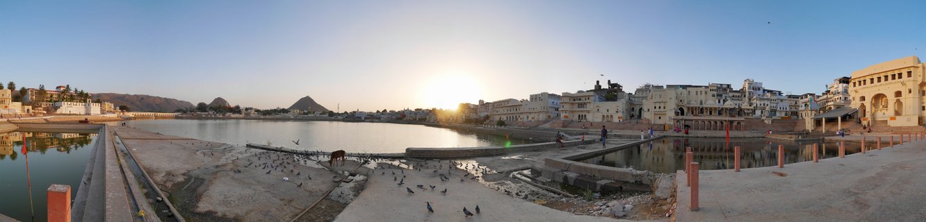 Panoramic view of Pushkar Lake in Rajasthan, India at sunset showing sacred water, stone ghats, traditional cream-colored buildings, pilgrims walking along the waterfront, cows, and hills in the background under golden sky