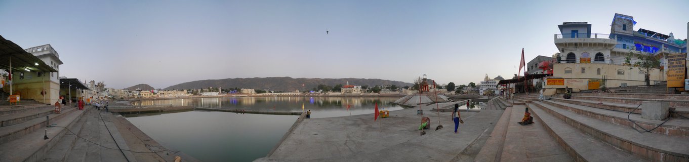 Wide panoramic view of Pushkar Lake in Rajasthan showing stone ghats with people walking, Hindu temple buildings, calm water reflecting the sky, and hills in the distance during evening light
