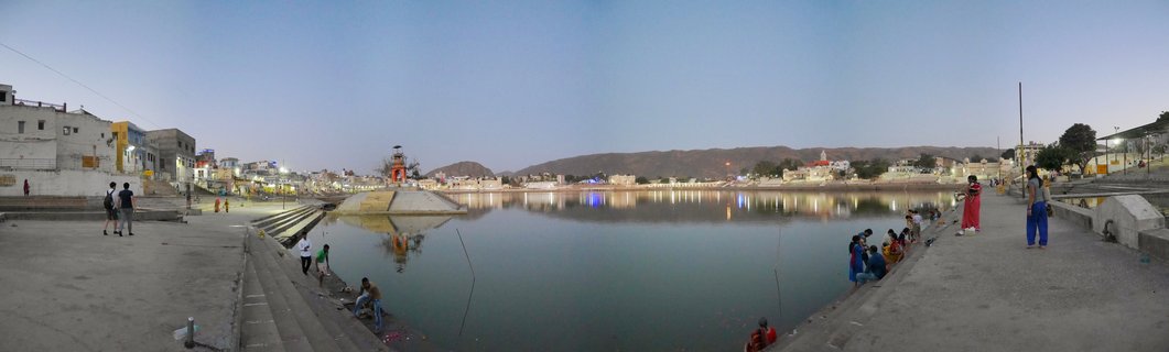 Panoramic view of Pushkar Lake at twilight showing pilgrims sitting on stone steps around the sacred water, with temples and buildings along the shoreline and hills in the background under a blue evening sky