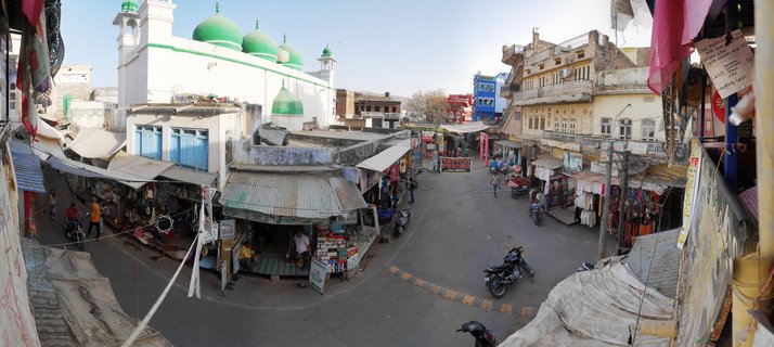 Panoramic street view of Pushkar market with white mosque featuring green domes, shops with awnings, motorcycles, and pedestrians on a clear day