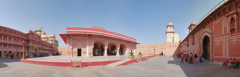 Wide courtyard of City Palace in Jaipur showing pink sandstone buildings with arched doorways, clock tower, and visitors walking on stone pavement under blue sky