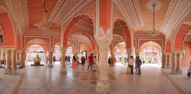 Interior view of ornate palace hall with pink walls, white decorative patterns, marble columns, arched doorways, chandeliers, and visitors walking on marble floor