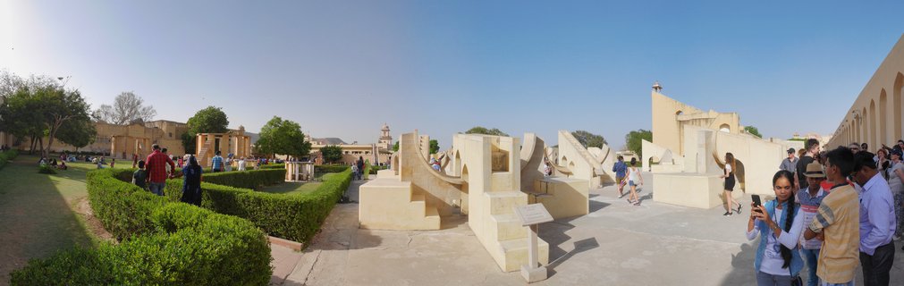 Wide view of Jantar Mantar observatory showing large beige stone astronomical instruments, green garden hedges, and tourists walking on paved courtyards under clear blue sky