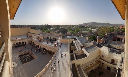 High-angle view of City Palace Jaipur showing multiple sandstone courtyards with tourists walking on terraces, domed pavilions, arched colonnades, and distant hills under bright afternoon sun