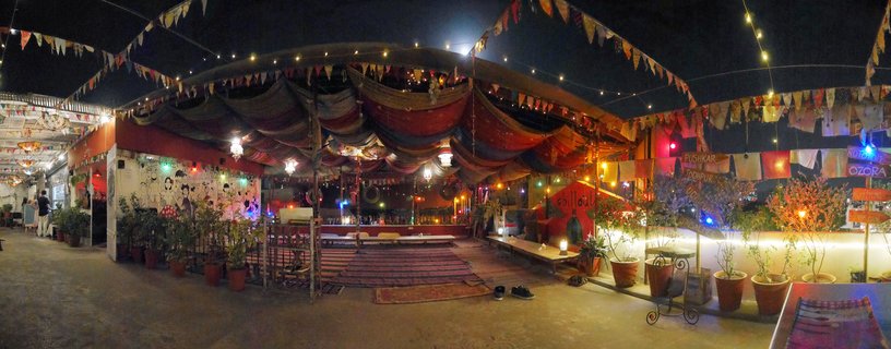 Nighttime view of an outdoor rooftop restaurant terrace with colorful fabric drapes overhead, string lights, potted plants, low seating with rugs, and directional signs pointing to Indian cities