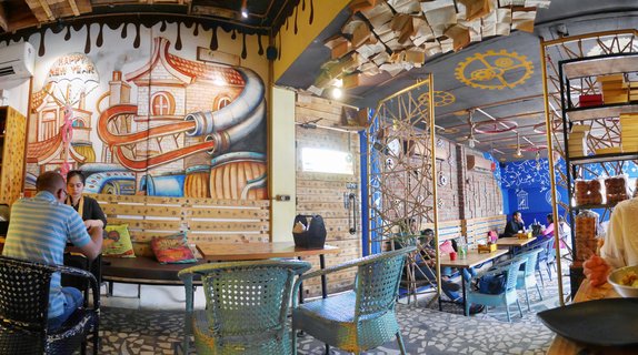 Interior of a themed cafe with books hanging from ceiling, colorful mural on wall, wooden tables, teal metal chairs, and patrons dining