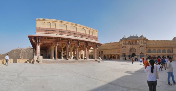 Wide courtyard at Amber Fort in Jaipur showing the ornate Diwan-i-Aam hall with sandstone columns on the left and palace buildings on the right, with tourists walking across the stone plaza under a clear blue sky