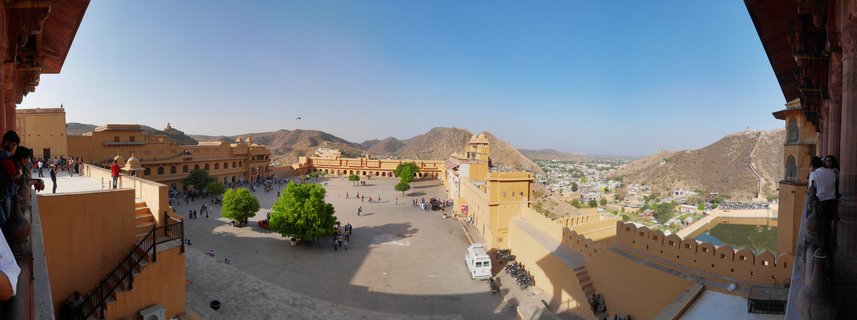 Wide view of Amber Fort courtyard in Jaipur showing golden sandstone buildings, tourists walking on stone pavement, green trees, and brown hills under clear blue sky