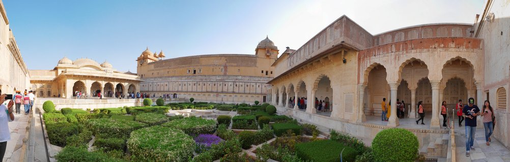 Panoramic view of a palace courtyard featuring formal gardens with trimmed hedges, surrounded by sandstone buildings with arched colonnades and domed pavilions under clear blue sky with tourists walking on stone walkways