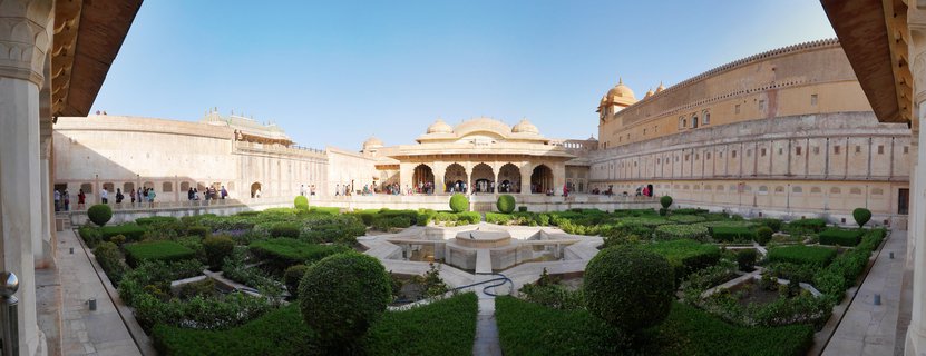 Panoramic view of Amber Fort courtyard showing formal garden with geometric hedge patterns, central stone fountain, sandstone palace buildings with domes and arched pavilions, tourists walking along walkways, clear blue sky