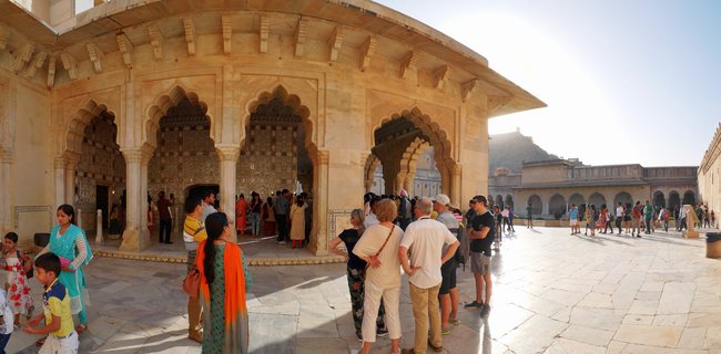 Tourists standing in the sunlit courtyard of Amber Fort palace in Jaipur, with ornate sandstone arches and carved doorways visible, hill fortress in background under clear sky