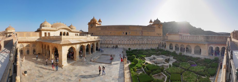 Panoramic view of Amber Fort courtyard in Jaipur showing tourists walking on marble pavement, Mughal garden with geometric hedges and fountain, sandstone palace buildings with domes and arches under clear blue sky