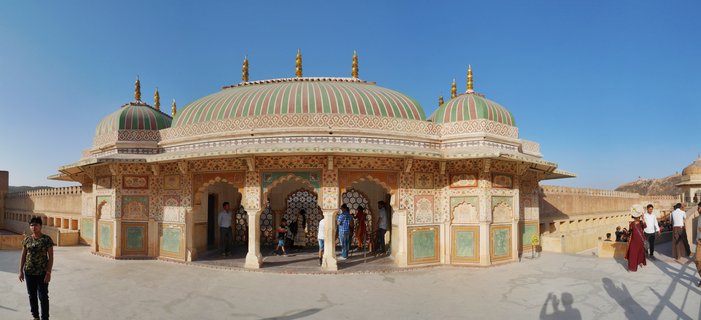 Ornate Rajput pavilion with three striped green and pink domes topped with golden finials, featuring arched doorways with decorative lattice screens, surrounded by tourists on a stone courtyard at Amber Fort, Jaipur, under clear blue sky