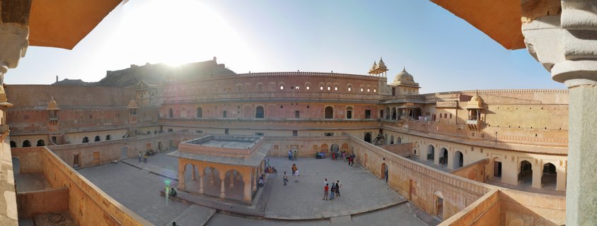 Aerial view of Amber Fort courtyard in Jaipur showing tourists walking on stone plaza surrounded by multi-level pink sandstone palace buildings with arched windows and ornate pavilions under bright sun and blue sky