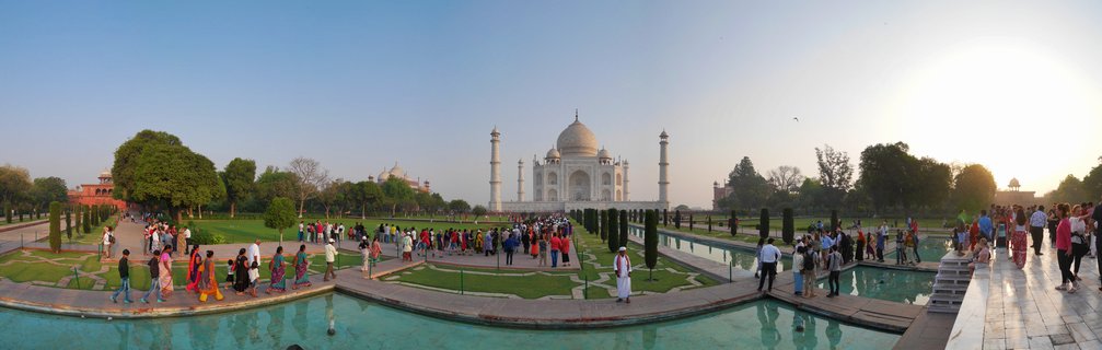 White marble Taj Mahal mausoleum with central dome and four minarets, surrounded by green gardens, reflecting pool in foreground, tourists walking on pathways, clear blue sky at sunrise, Agra India