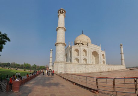 The Taj Mahal white marble mausoleum with its iconic central dome and four minarets stands against a clear blue sky in Agra, India. Tourists walk along the red sandstone pathway in the foreground, with manicured green lawns visible to the left. The morning sunlight illuminates the intricate architectural details of this UNESCO World Heritage Site.