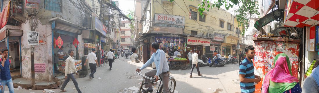 Street scene in Delhi showing people walking, a man riding a bicycle, shop storefronts with signs, overhead electrical wires, and buildings lining a narrow commercial street