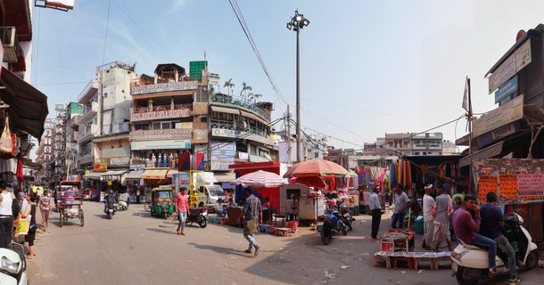 Busy street market in Delhi with colorful vendor stalls, auto-rickshaws, motorcycles, pedestrians, and multi-story buildings with restaurant signs under a clear blue sky