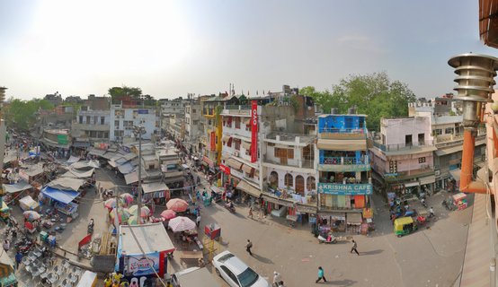 High-angle view of a busy Delhi street market showing colorful vendor umbrellas, auto-rickshaws, pedestrians, and multi-story buildings with commercial signage including Krishna Cafe and OYO hotel