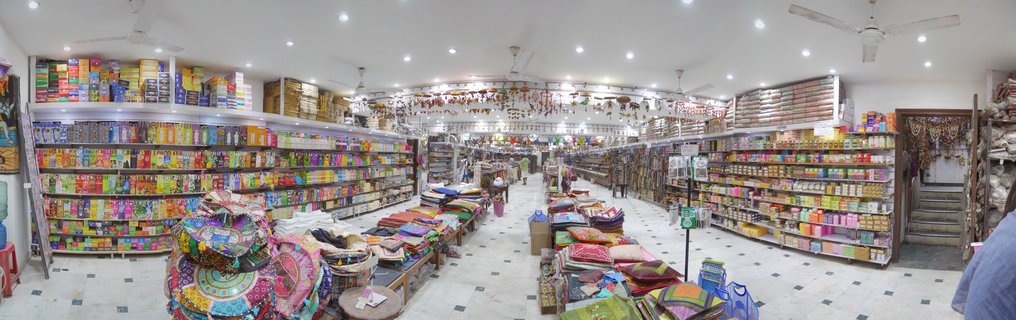 Interior view of a textile shop in Delhi with shelves of colorful fabrics, stacks of embroidered cushions on tables, ceiling fans, and bright overhead lighting