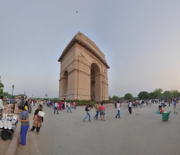 A large sandstone arch monument with crowds of people walking on a paved plaza in front, street vendors on the left side, trees visible in the background under a hazy sky