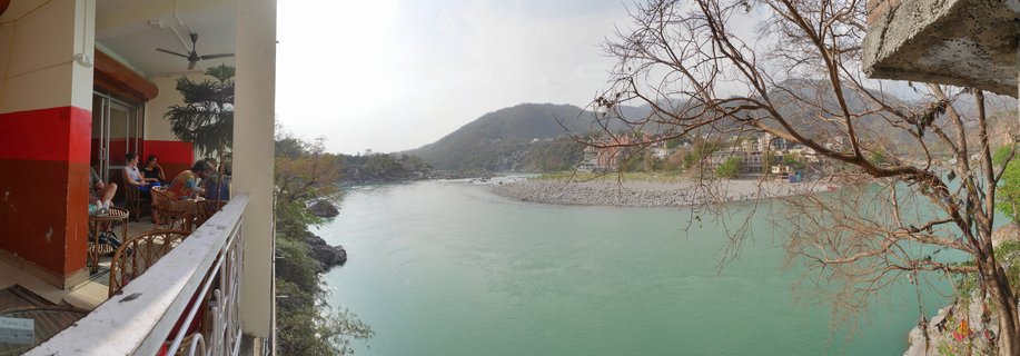 View from a cafe terrace showing people seated at tables on the left, overlooking a wide turquoise river with rocky banks, buildings, and forested hills in Rishikesh, India