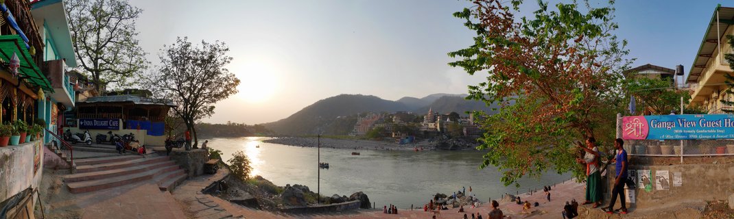 Panoramic view of the Ganges River in Rishikesh, India at sunset with people on stone steps, buildings along the riverbank, boats on the water, and mountains in the background