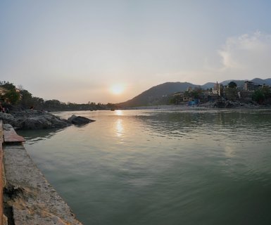 Sunset view over the Ganges River in Rishikesh with golden sun reflection on water, buildings and ashrams on the far bank, and mountains in the background