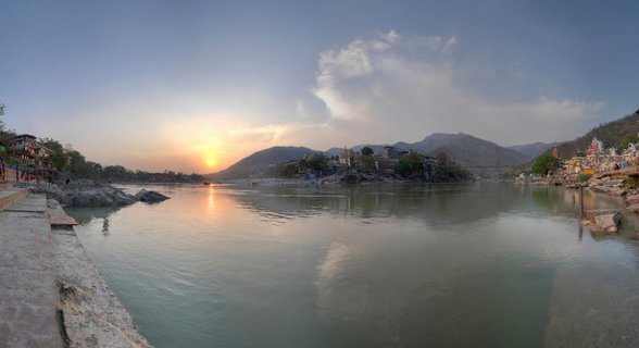 Wide panoramic view of the Ganges River at sunset in Rishikesh, India, with temples, ghats, and mountains visible along the riverbanks under a golden sky.