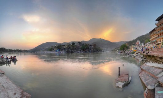 Wide panoramic view of the Ganges River in Rishikesh, India at sunset with people sitting on stone steps, buildings along the shore, and mountains in the background